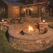 EP Henry Rustic, outdoor evening scene with lit fire pit. Small curved wall borders the pation and a table and chairs can be seen in the background. Cast Stone Wall Face Shells Breckenridge, Double Sided Cast Stone Wall Breckenridge, Chapeau 16 Caps Brownstone